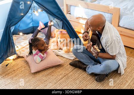 African American father and daughter playing guitar in bedroom play tent with lights and teddy bear Stock Photo