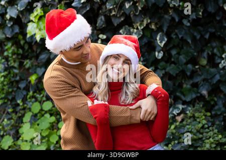 Diverse couple hugging and smiling while wearing Santa hats and knit sweaters in garden Stock Photo