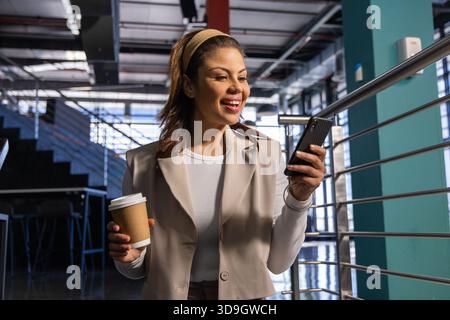 Mid adult woman wearing beige blazer standing in office holding coffee cup and checking smartphone Stock Photo