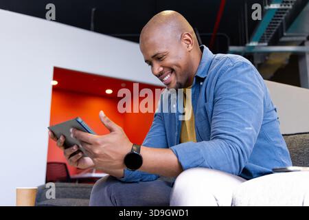 African American man using tablet on gray sofa in office lounge checking smartwatch by coffee cup Stock Photo