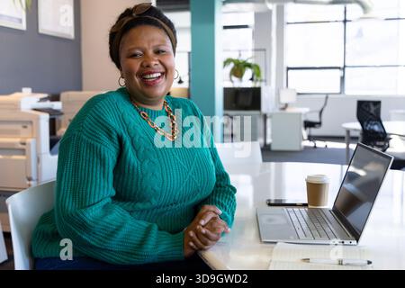 African American woman sitting at modern office desk smiling and working on laptop with coffee cup Stock Photo