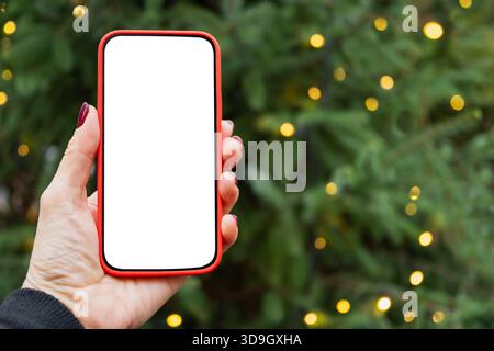 Smartphone with empty blank screen in womans hand against Christmas tree with light garland. Screen display for Festive Xmas Holiday Background. Space Stock Photo