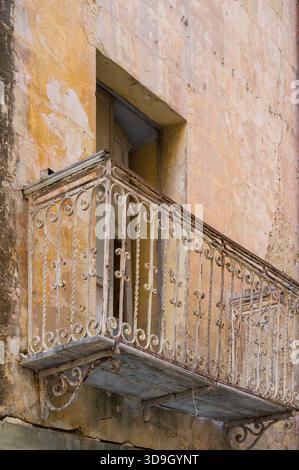 A weathered balcony with ornate ironwork clings to a crumbling facade. The peeling paint and rusted metal hint at a bygone era, evoking a sense of fad Stock Photo