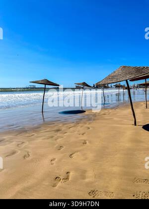 Empty beach parasols on wet sand along the sunny Essaouira shoreline in Morocco, gentle waves rolling in under the blue midday sky Stock Photo