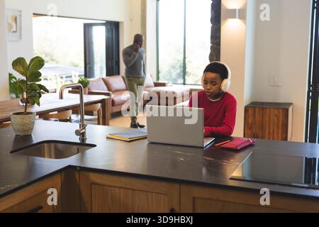 African American boy with headphones working laptop at kitchen island, copy space Stock Photo