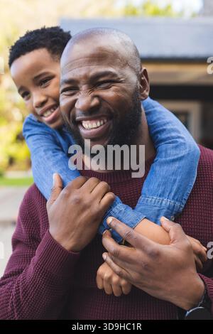 African American father carrying son wearing blue shirt on back outside home displaying smartwatch Stock Photo