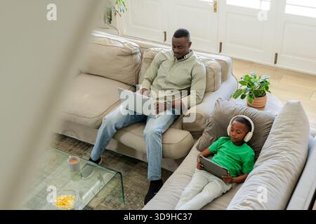 African American father and child sitting on beige sectional at home using laptop and tablet Stock Photo