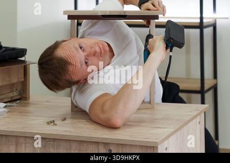 Young man in white shirt lies awkwardly under light wood frame on table, using blue drill to screw from below with tools scattered. Stock Photo