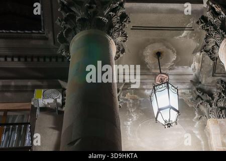 Single ornate column with Corinthian capital and hanging lantern next to modern air conditioner on aged wall at night Stock Photo