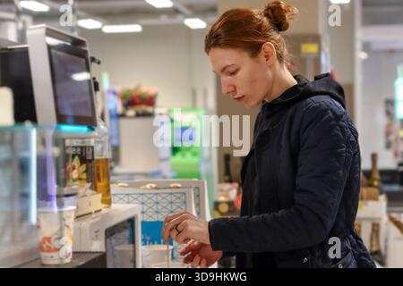 Young red-haired woman in dark attire selects additive for her coffee at supermarket beverage counter. Stock Photo