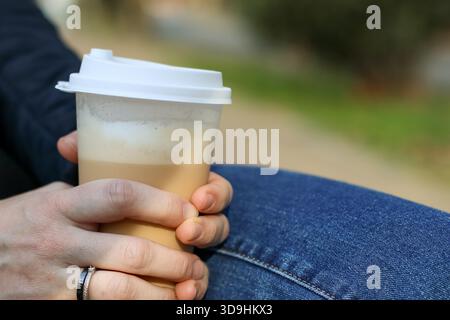 Legs in jeans on wooden park bench with hand grasping takeaway beverage and smartphone lying nearby Stock Photo