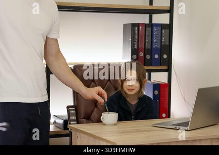 An adult's arm uses a spoon to stir a white mug placed on the desk as a preteen girl looks on with mild interest beside her open laptop. Stock Photo