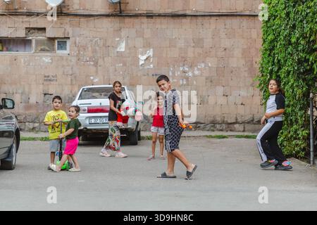Vayk, Armenia - 16 August 2025: View of children playing in the street near a light-colored car against the backdrop of a weathered brick wall. Stock Photo