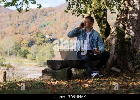 A young man sits on a wooden bench under a mossy tree, talking on his phone while holding a coffee cup next to his open laptop in an autumn mountain l Stock Photo
