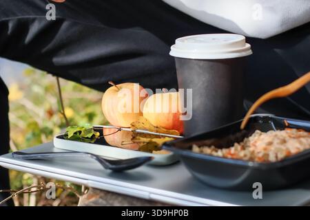 Wide view of tray holding smartphone, spoon, apples, coffee cup, and black container filled with rice, vegetables, and fork in natural setting. Stock Photo