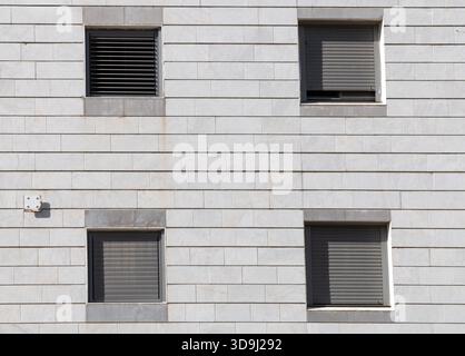 Symmetrical close view of a modern building facade with square windows and closed roller shutters on light stone tiles, showing geometric pattern, minimal lines, and urban housing architecture Stock Photo