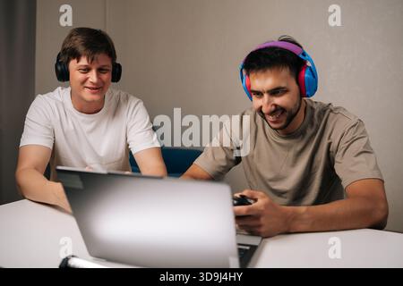 Portrait of two cheerful young men gathered in living room, laughing and focused on laptop and controllers while gaming together, showcasing Stock Photo