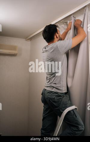 Strong man standing on a stepladder, busy installing new curtains on rail, performing household chores and home maintenance, focusing on domestic Stock Photo