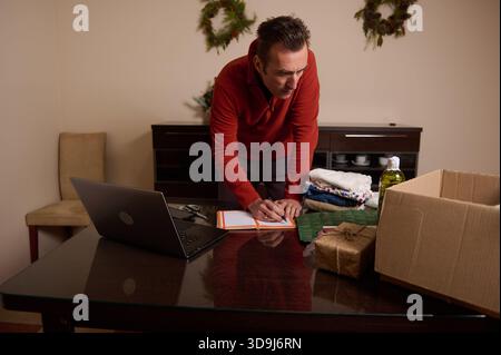 A man in a red sweater organizes papers and gifts on a dining room table, readying packages for shipment amid holiday decor and a laptop. Stock Photo