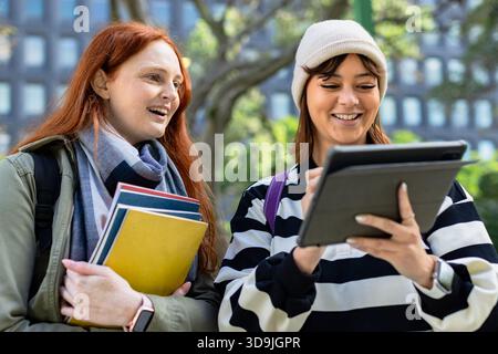 Two female students collaborating on campus walkway, holding notebooks and using stylus tablet Stock Photo