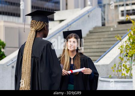 Diverse female friends wearing gowns caps conversing on campus holding diploma with red ribbon Stock Photo