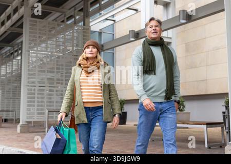 Senior couple walking together along brick promenade under glass canopy holding paper shopping bags Stock Photo