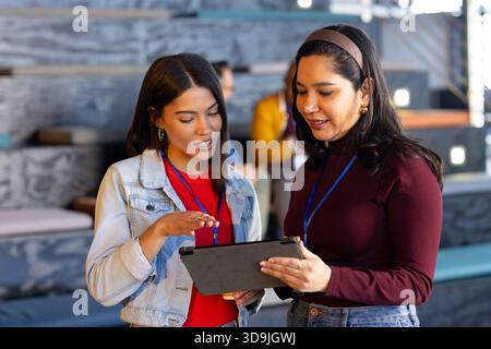 Diverse female colleagues standing in conference lounge holding tablet and wearing badge lanyards Stock Photo