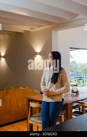 Asian woman standing in bright living dining area holding ceramic mug near leather sofa, copy space Stock Photo