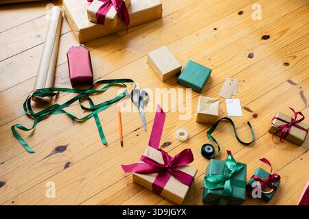 Gift box is sitting on wooden tabletop, kraft paper rolls and ribbon spools are lying nearby Stock Photo