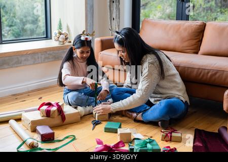 Indian mother and daughter sitting on wooden floor at home wrapping holiday gift boxes with ribbons Stock Photo
