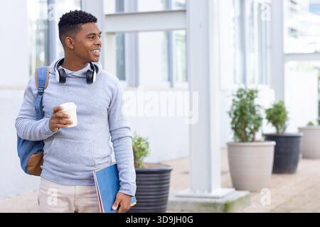African American man walking on campus wearing headphones carrying backpack holding coffee and file Stock Photo