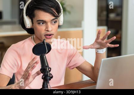 Asian man speaking into condenser microphone wearing headphones, gesturing at wooden desk in studio Stock Photo