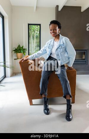 African American woman perching atop brown leather armchair in modern living room with fireplace Stock Photo