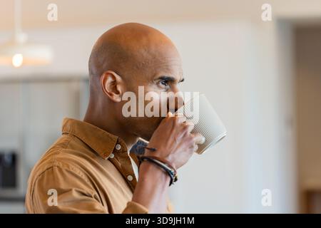 Adult African American man sipping from white mug wearing brown shirt with bracelets in kitchen Stock Photo