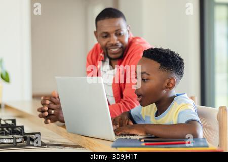 African American father and son leaning over home kitchen island using laptop and colored pencils Stock Photo