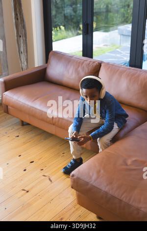 African American boy sitting on sofa at home wearing headphones using smartphone, copy space Stock Photo