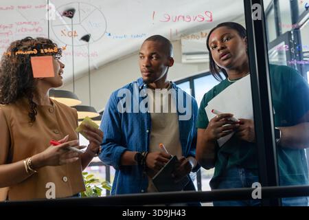 Diverse coworkers brainstorming at glass whiteboard in modern office using sticky notes and markers Stock Photo