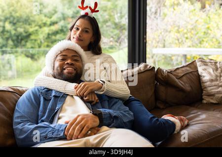 Diverse couple hugging on brown leather sofa by large window wearing antler headband, Santa hat Stock Photo
