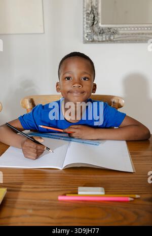 African American child sitting at home table wearing blue tee writing in notebook holding pencil Stock Photo