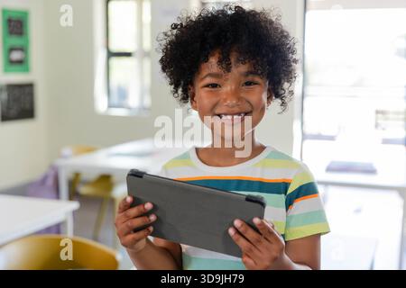 African American child in striped shirt holding tablet with dark case and smiling in classroom Stock Photo
