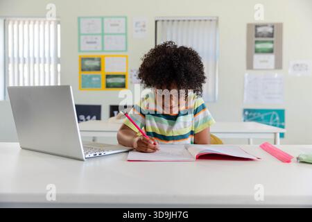 African American youth in striped tee sitting at classroom desk writing in pink notebook by laptop Stock Photo
