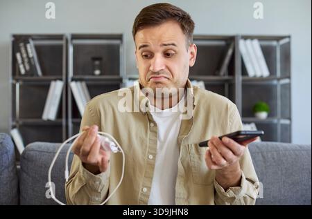 Upset frustrated man holding broken mobile phone and charger with cable in hands Stock Photo