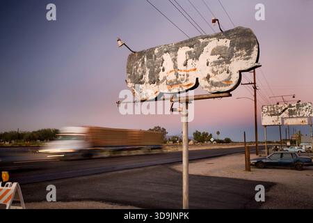 A worn out cafe sign stands on a rusty pole as trucks drive by at dusk Stock Photo