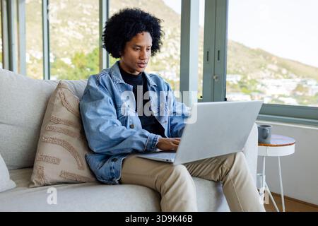 African American man typing on laptop on sofa with coffee mug in living room overlooking hillside Stock Photo
