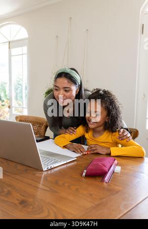 Mother and daughter leaning over dining table at home using laptop writing in notebook with pencil Stock Photo