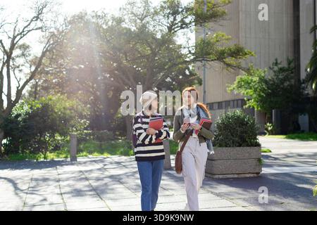 Diverse female students walking on campus walkway chatting while holding notebooks and smartphone Stock Photo