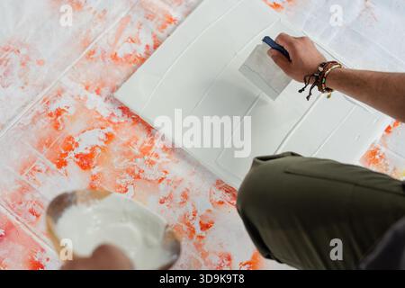 Indian man kneeling in workshop on splattered cloth troweling white plaster onto panel, copy space Stock Photo