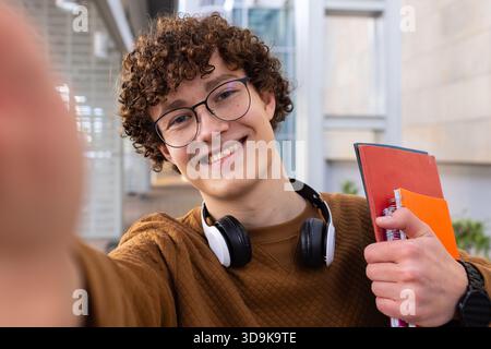 Male student holding notebooks and smartphone taking selfie in university corridor with headphones Stock Photo
