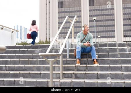 Mature adult man sitting on steps by handrail at plaza wearing headphones and satchel, copy space Stock Photo