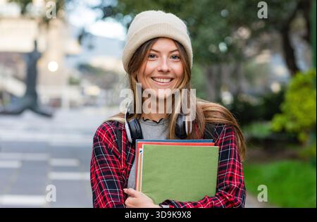 Female student standing on campus walkway wearing plaid shirt holding notebooks and headphones Stock Photo